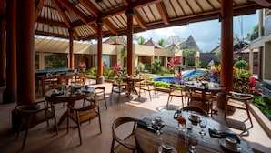 Un espace extérieur abrité, aménagé pour les repas, avec des tables et des chaises en bois, une piscine et des plantes tropicales.
