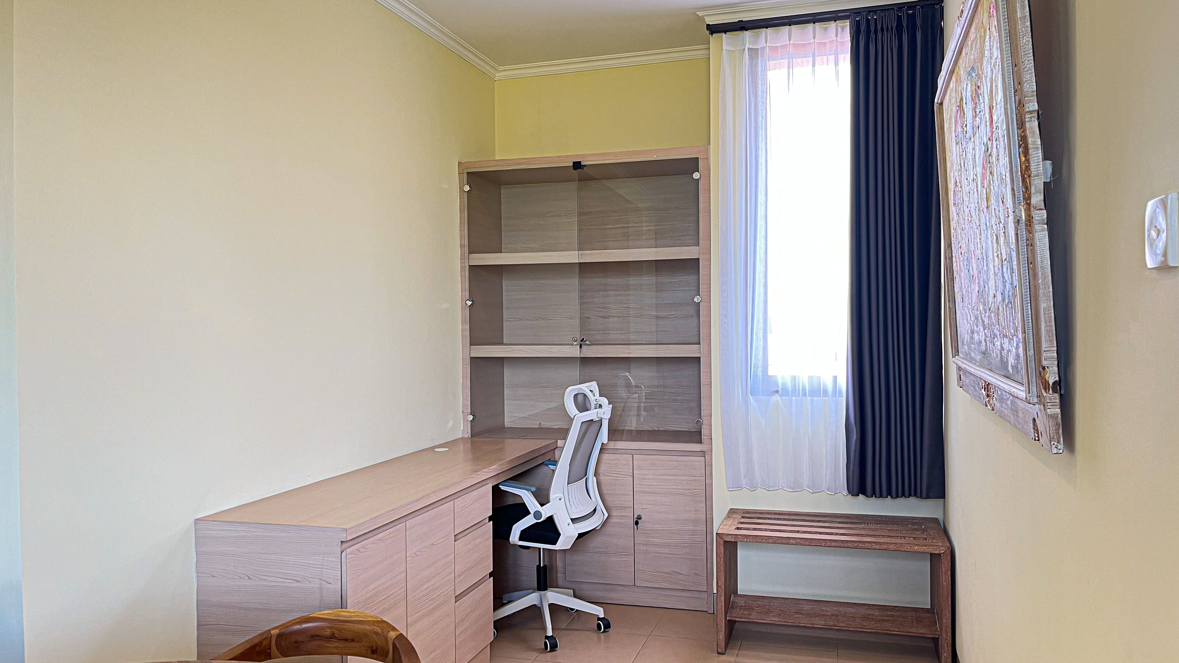A room with a wooden desk, a white office chair, a small wooden bench, and a large window with curtains.