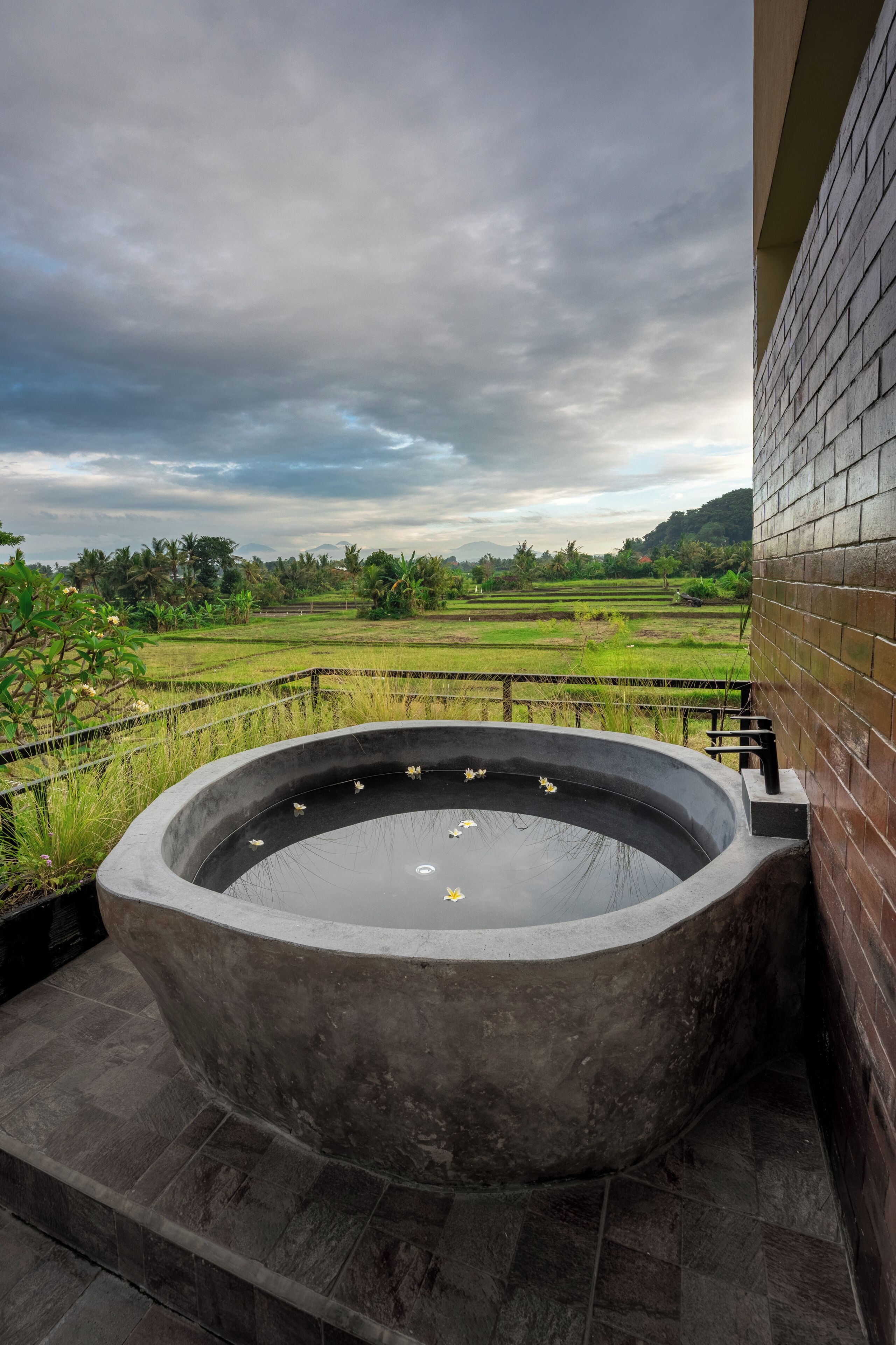 A concrete hot tub on a tiled patio with a view of a green landscape and a cloudy sky.