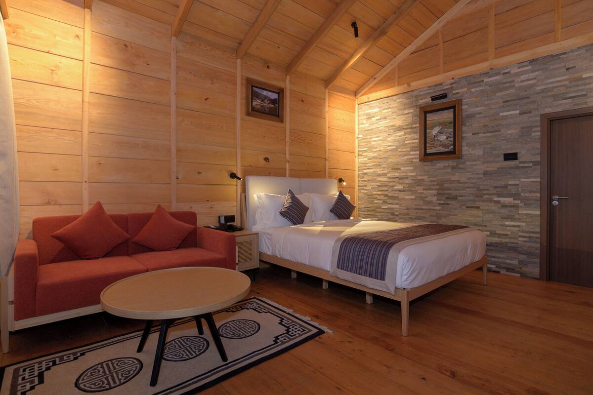 Una habitación acogedora con techo de madera, una cama con almohadas, un sofá rojo, una pequeña mesa y una pared de piedra.