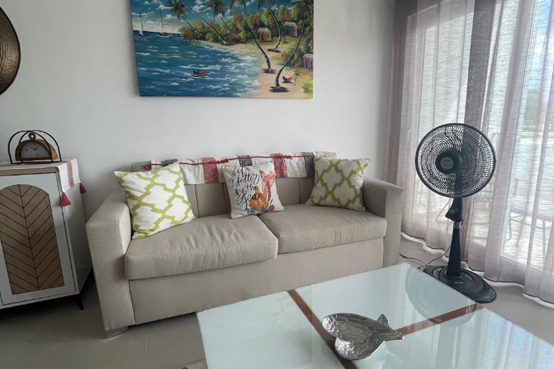 Un salon avec un canapé beige, des coussins à motifs, une table basse en verre, un ventilateur fixé au mur et un tableau représentant une scène de plage.