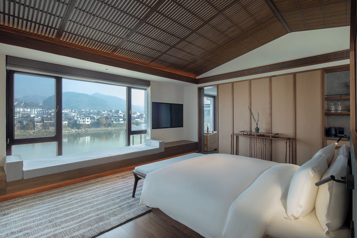 Quarto com uma janela grande, uma cama, uma TV e vista para uma cidade e montanhas.