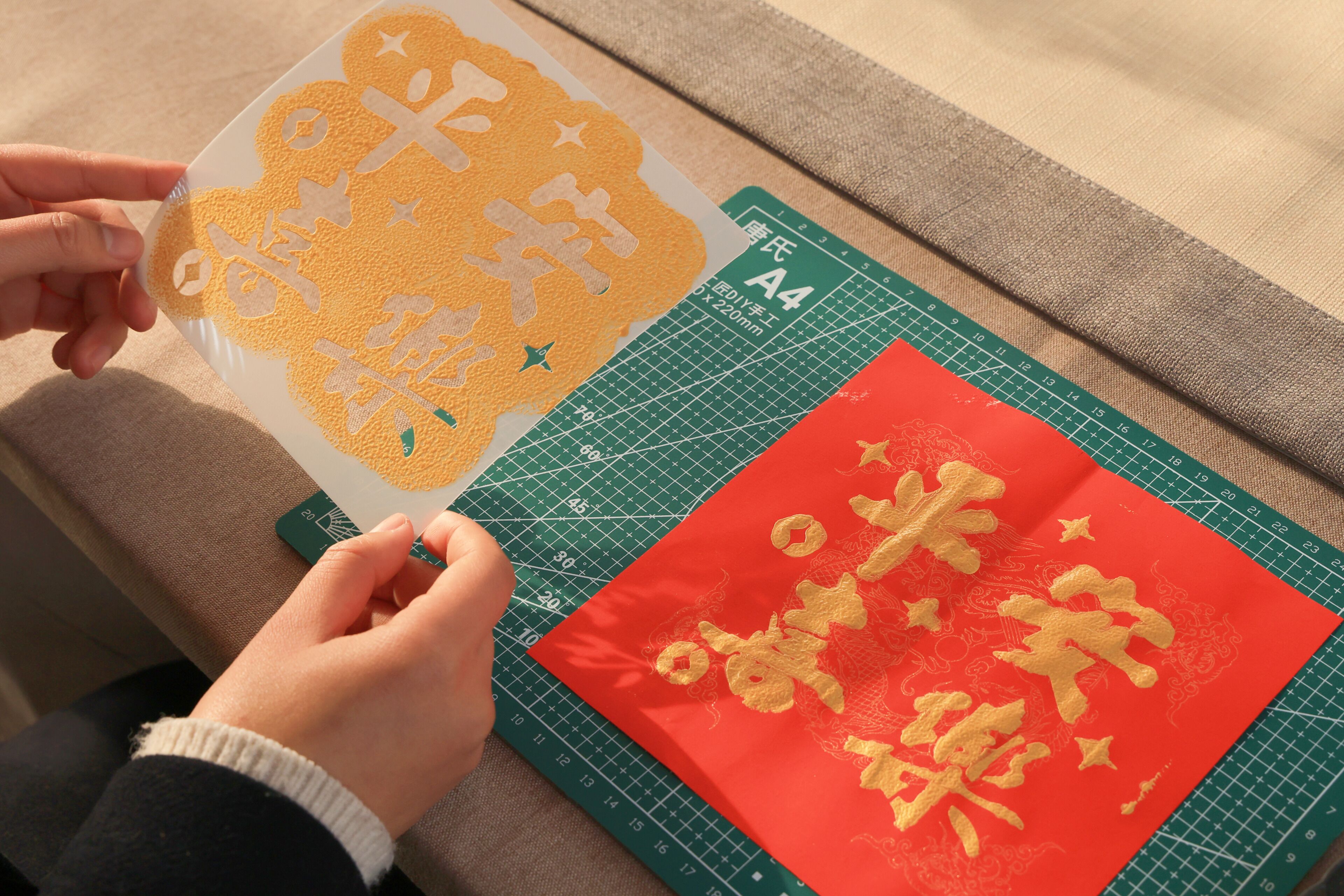A person using a cutting mat with a grid pattern and a red fabric with golden embroidery.