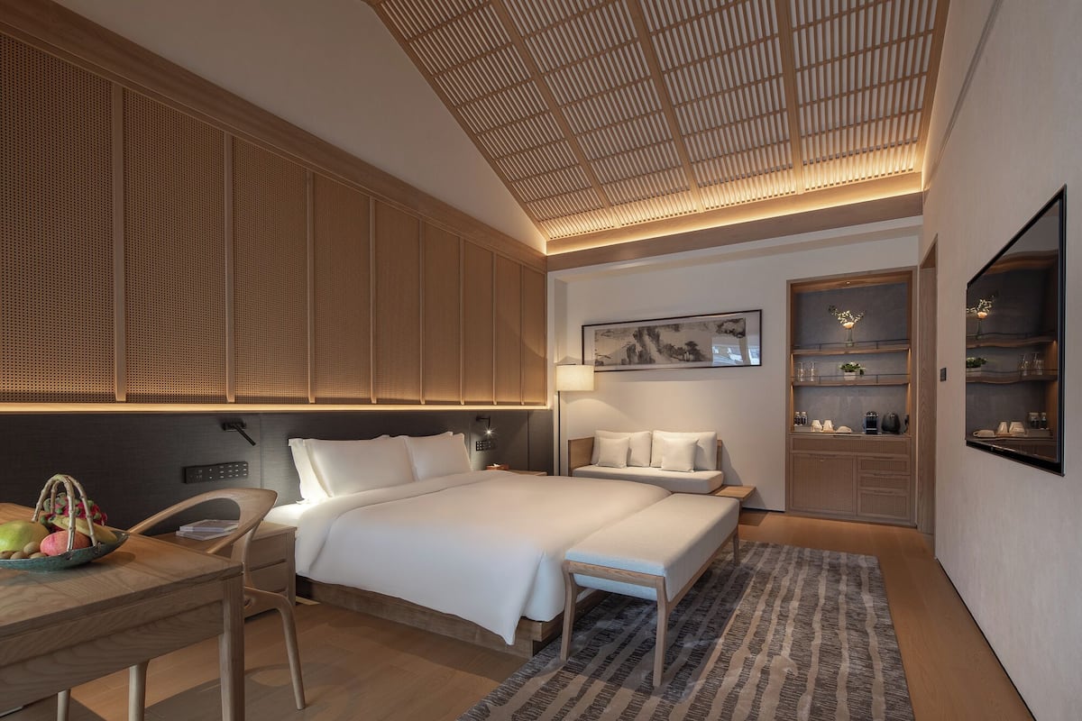Quarto moderno com uma cama grande, mesas de cabeceira, uma escrivaninha com uma cesta de frutas, uma prateleira na parede com objetos decorativos e um teto com design de ripas de madeira.