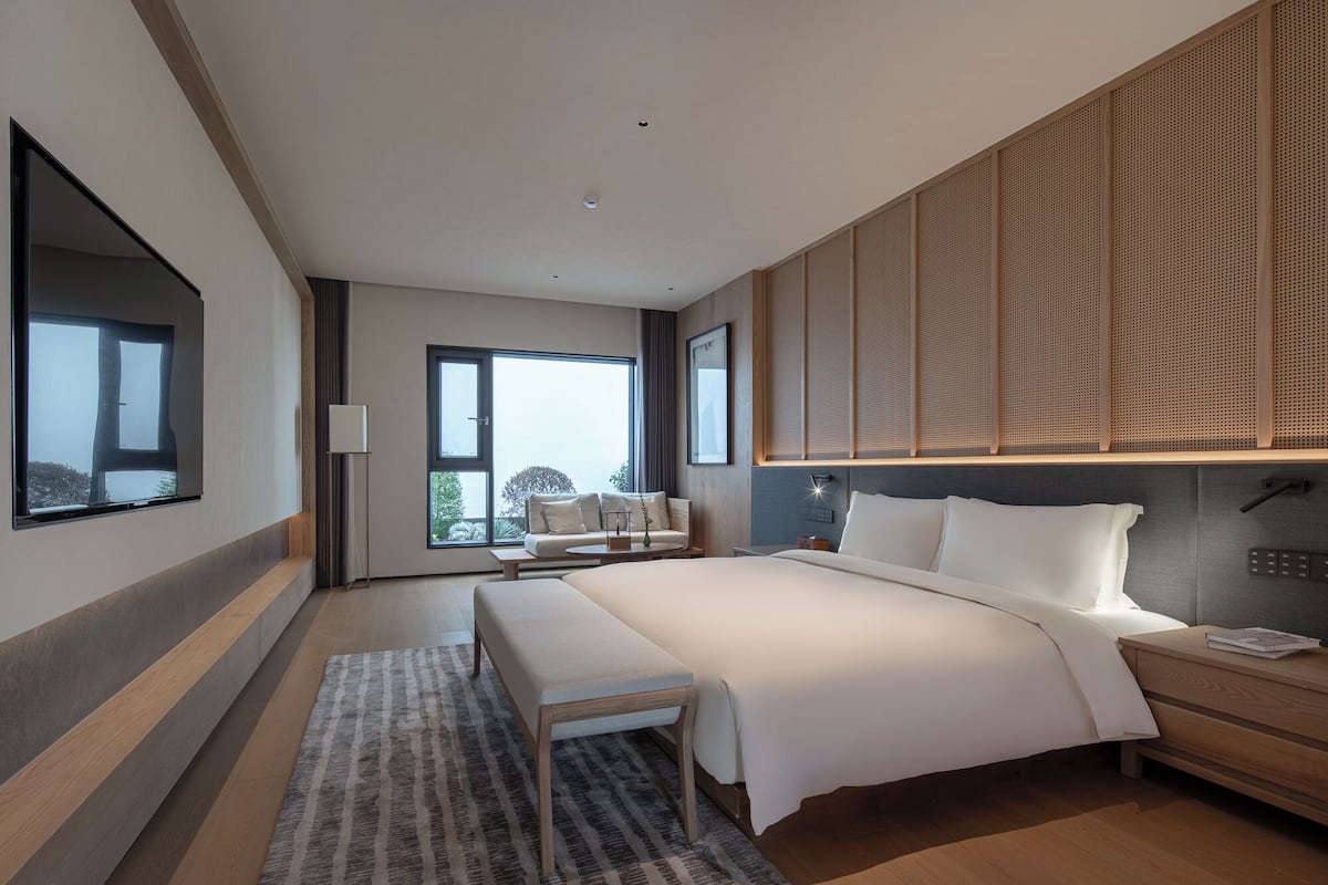 Quarto moderno com cama grande, mesa de cabeceira, um banco e vista para o exterior.