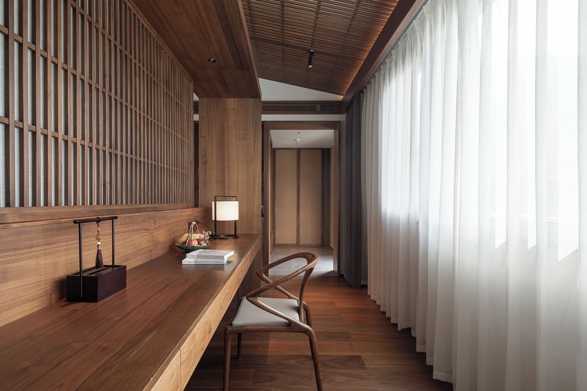 A modern hotel room with a wooden reception desk, a chair, a lamp, and a view through sheer curtains.