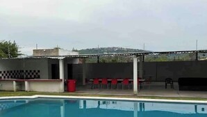 A swimming pool with a covered patio area, red chairs, and a barbecue grill. - Leon House (Jardines de la Calera)