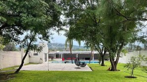 A backyard with a pool, a covered patio, and a view of a hilly landscape. - Leon House (Jardines de la Calera)