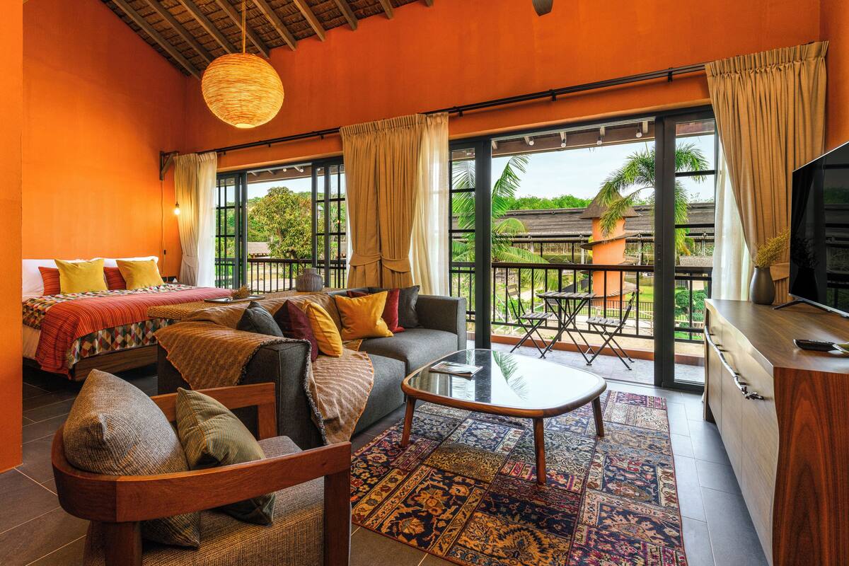 A room with a bed, sofa, and a dining area with a table and chairs. There is a balcony with a view of the outdoors.