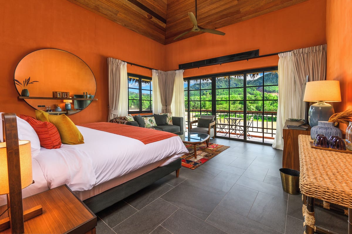 A bedroom with a large bed, a round mirror, a ceiling fan, and a view of lush greenery through large windows.