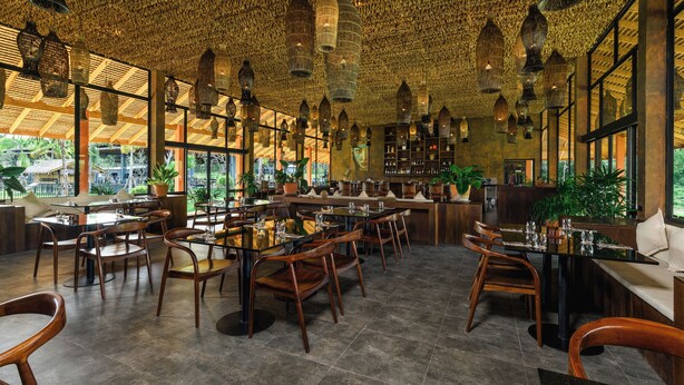 A restaurant with wooden tables and chairs, hanging birdcages, and large windows overlooking a garden. - Niran Farm (Si Sunthon)