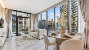 A modern living room with a sofa, coffee table, and dining table set for four. Large windows offer a view of the cityscape and construction cranes.