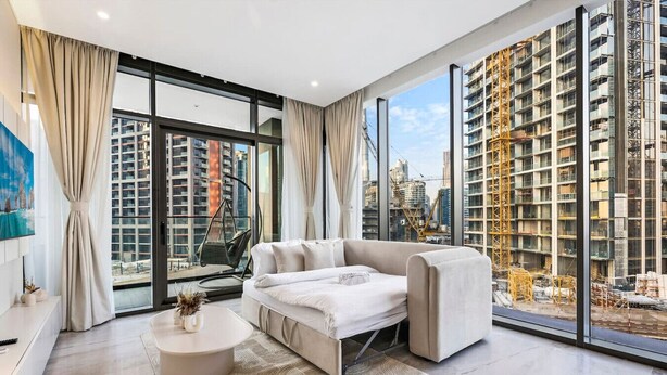 A modern living room with a sofa, a coffee table, and a view of a cityscape with buildings and construction cranes.