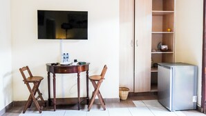 Ein kleines Zimmer mit an der Wand montiertem Fernseher, einem Holztisch mit zwei Klappstühlen, einem Minikühlschrank und einem Schrank mit Regalen.