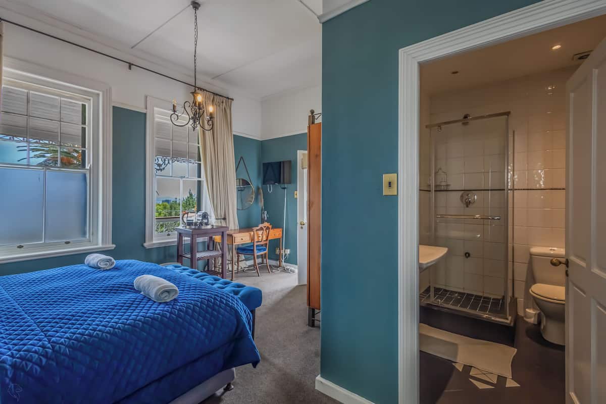 A bedroom with a large bed, a desk, and a chair, a bathroom with a shower, and a view of greenery outside.