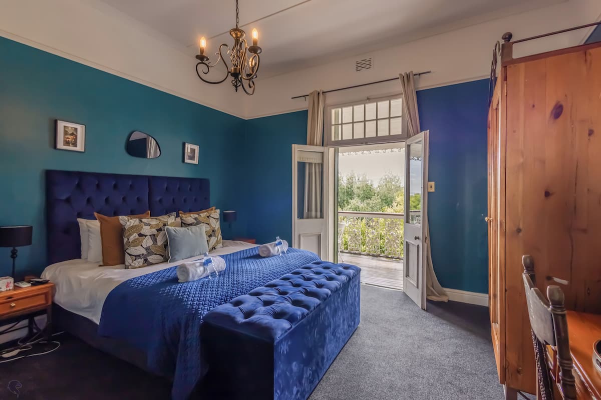 A bedroom with a large bed, a blue upholstered bench, a chandelier, and a view of the outdoors through a door.