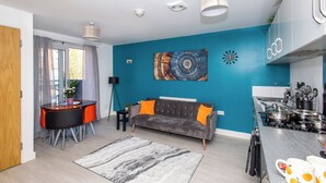 A modern living room with a grey sofa, orange cushions, a round dining table with chairs, a coffee table, and a kitchen area with a stove and cabinets. - Wakefield Westgate Station - 2 Bed 2 Bath (Wakefield)
