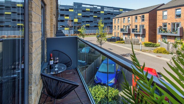 A balcony with a table and chairs, a blue car parked on the street, and a modern building in the background. - Wakefield Westgate Station - 2 Bed 2 Bath (Wakefield)