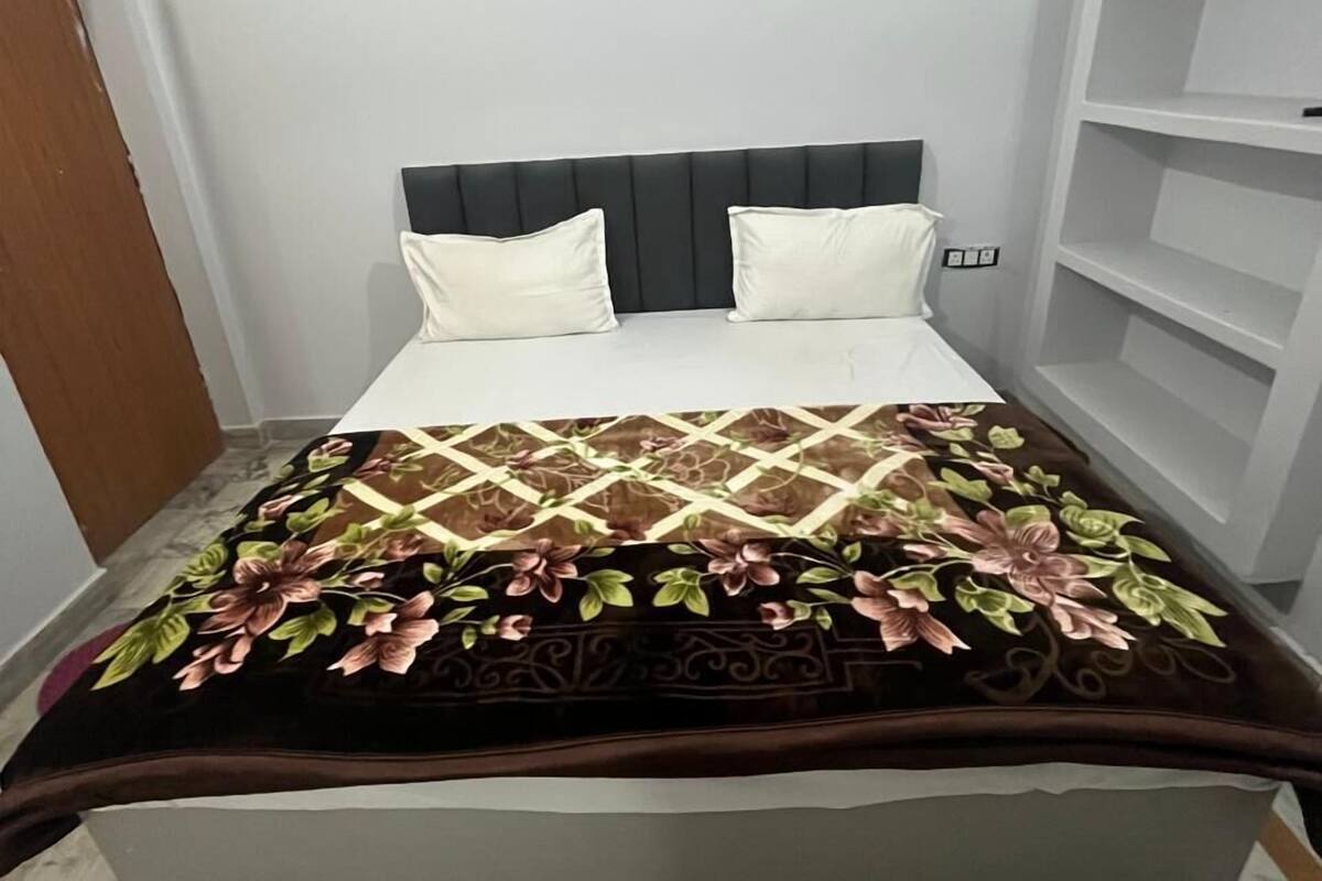 A bed with a dark headboard, white pillows, and a floral-patterned bedspread. There is a white shelving unit to the right.