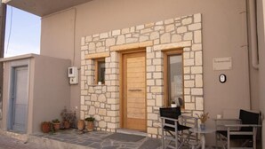 A stone facade building with a wooden door and two windows, a small outdoor seating area with a table and chairs, and potted plants on the ground.