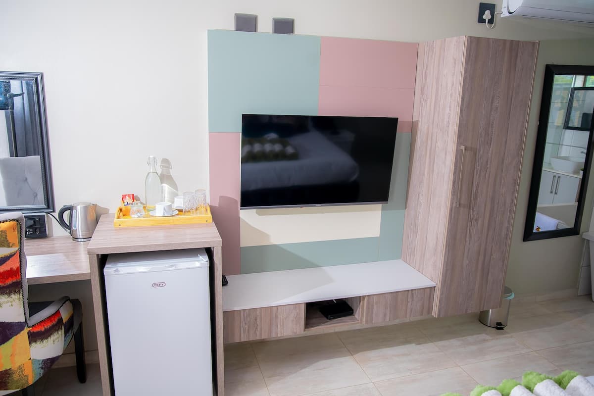 Habitación de hotel compacta con una televisión de pantalla plana montada en la pared, un pequeño rincón de kitchenette con refrigerador y una cama con cabecera.