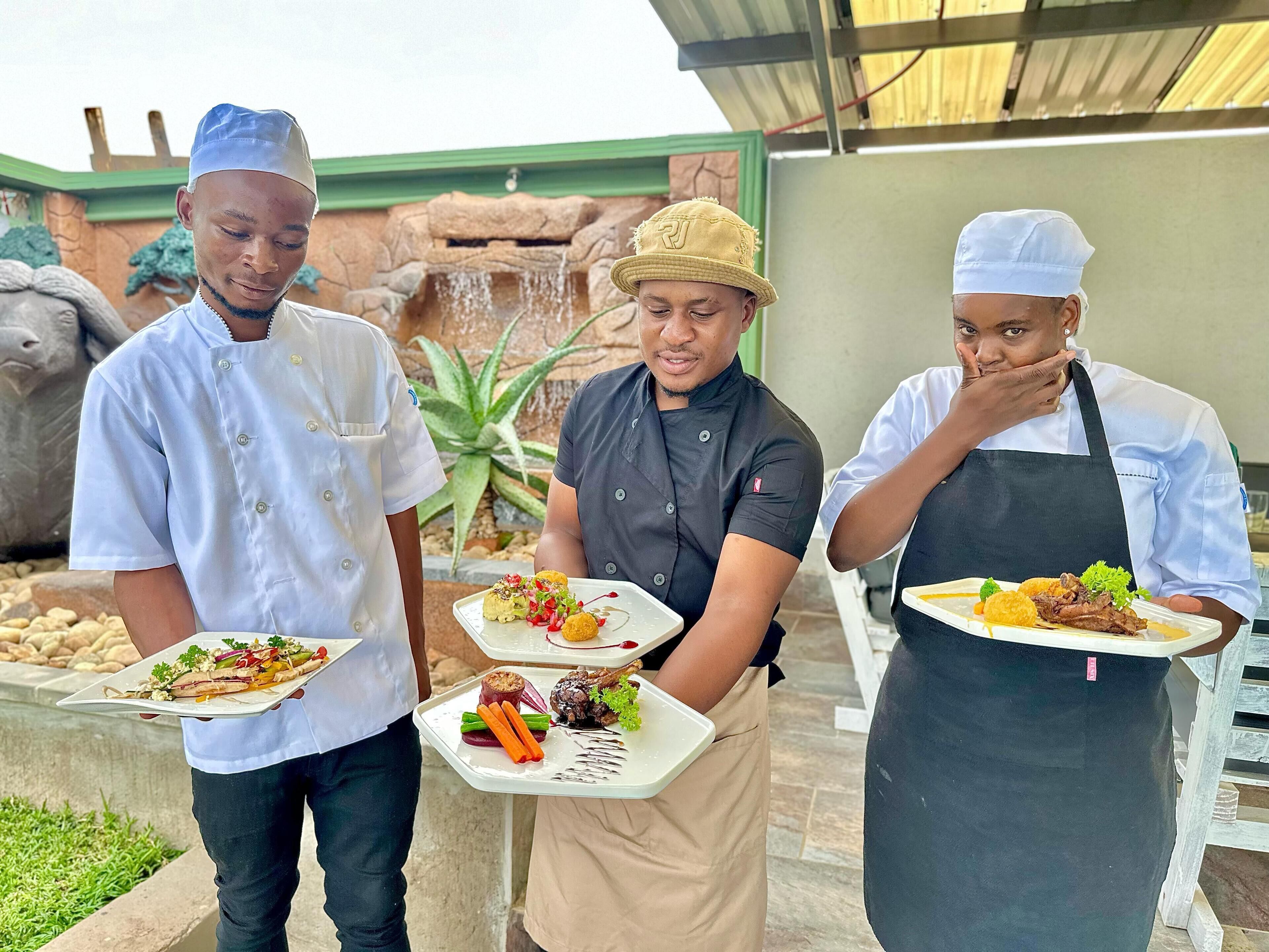 Three chefs holding plates of food in an outdoor setting with a waterfall and greenery in the background.