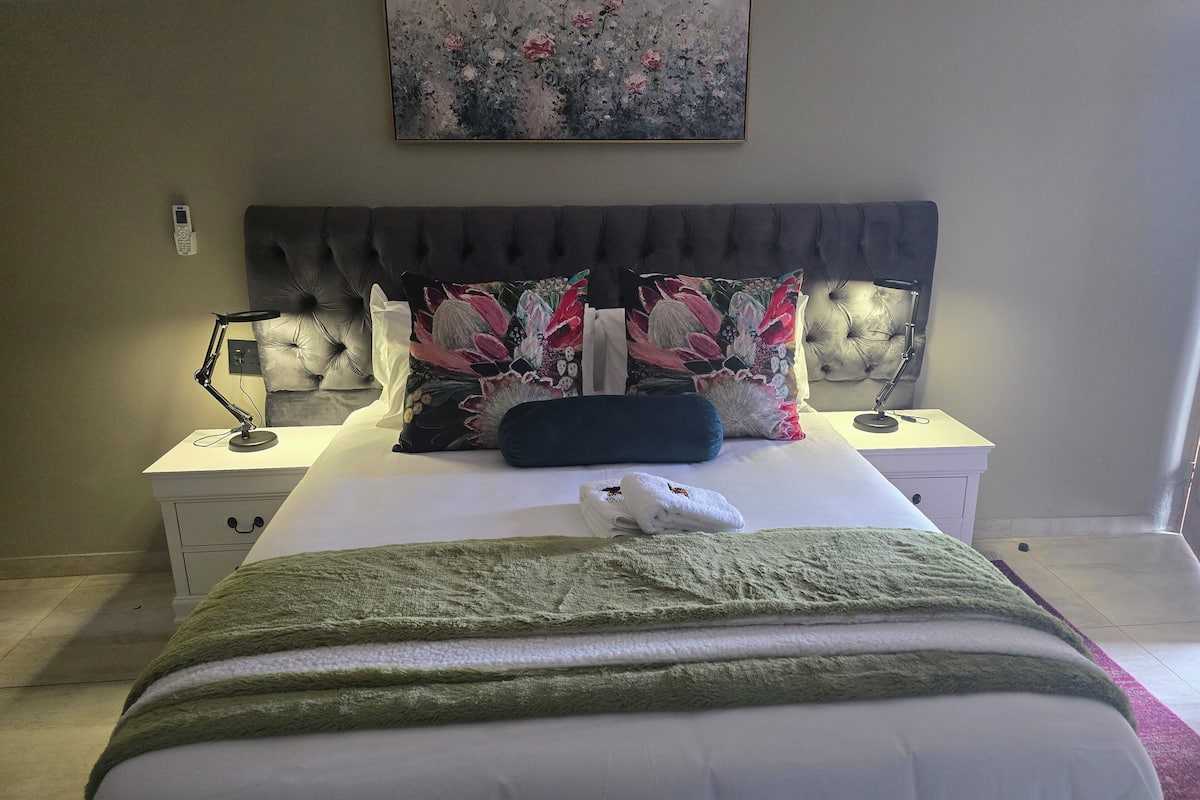 Una cama bien hecha con una manta verde, una almohada floral y una toalla blanca a un lado. Hay una lámpara de noche, una mesita auxiliar y un cuadro en la pared sobre la cama.