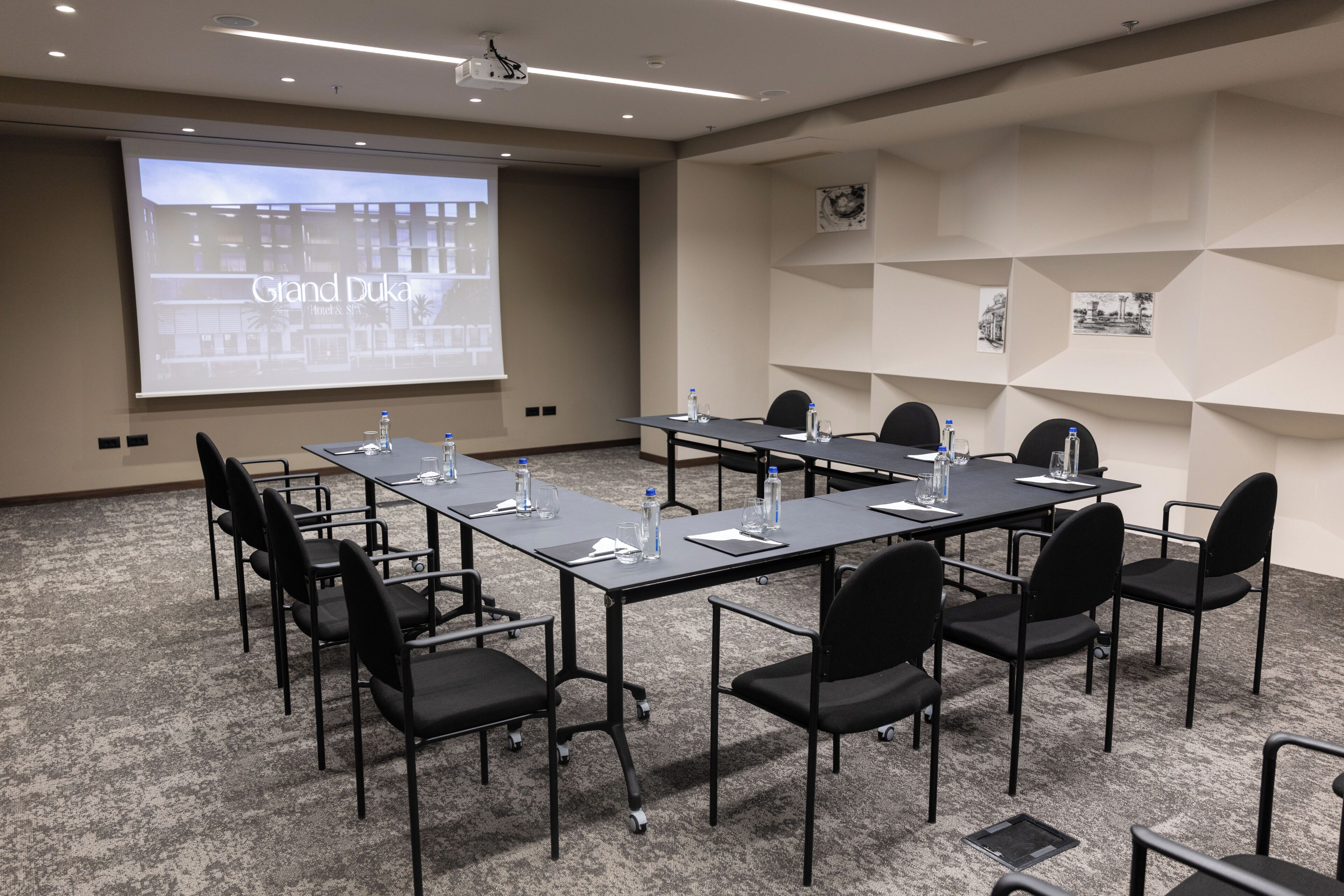 Une salle de conférence avec une longue table, des chaises noires, des bouteilles d’eau et un écran de projection sur lequel est affiché le titre « Grand Dula ».