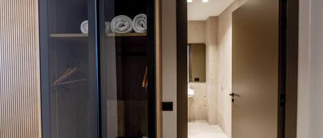 A hallway with a glass door leading to a room with a bed, a wooden floor, and a towel rack with rolled towels.