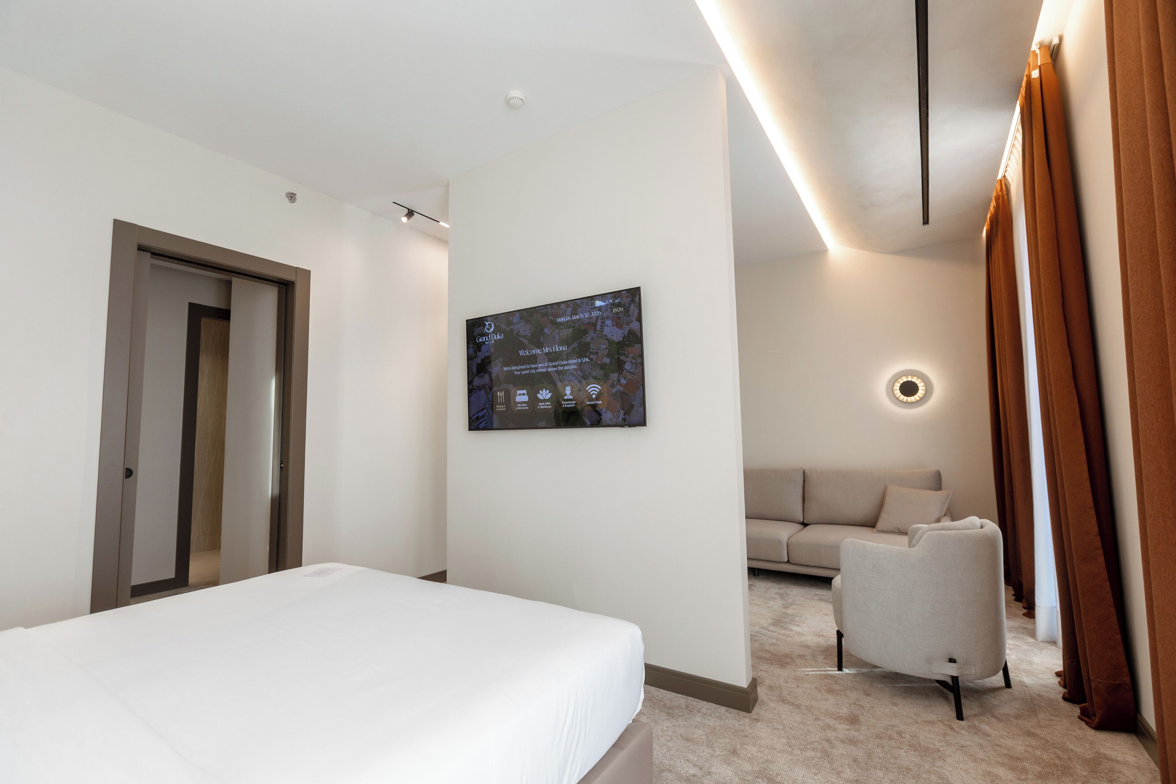 A modern hotel room with a bed, a sofa, a chair, a wall-mounted TV, and a door.