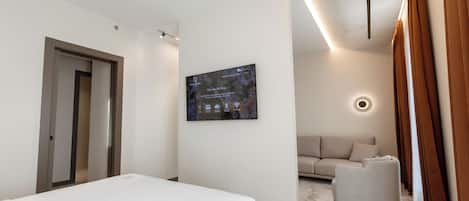 A modern hotel room with a bed, a sofa, a chair, a wall-mounted TV, and a door.