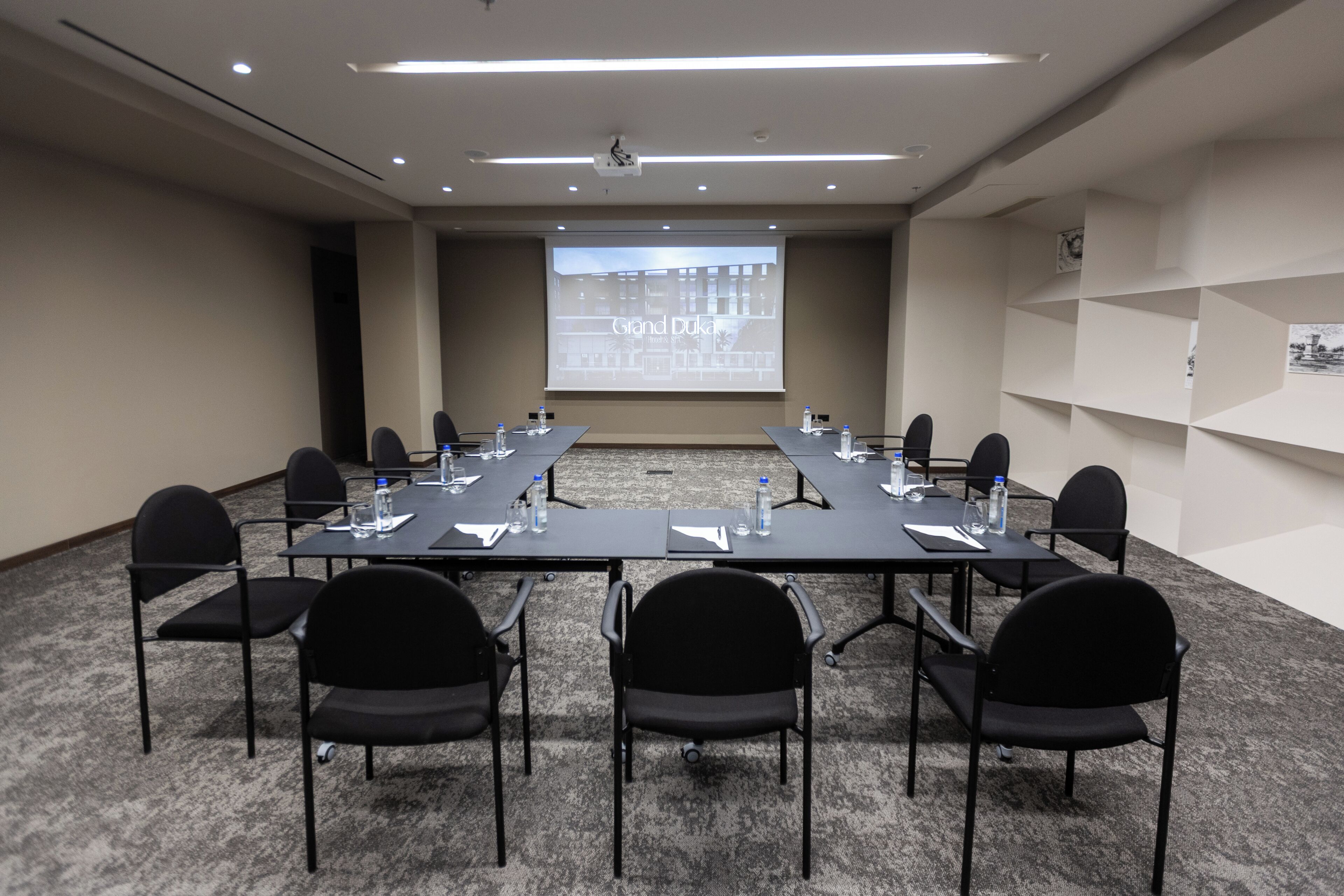 A conference room with a long table, black chairs, a projector screen, and a carpeted floor.