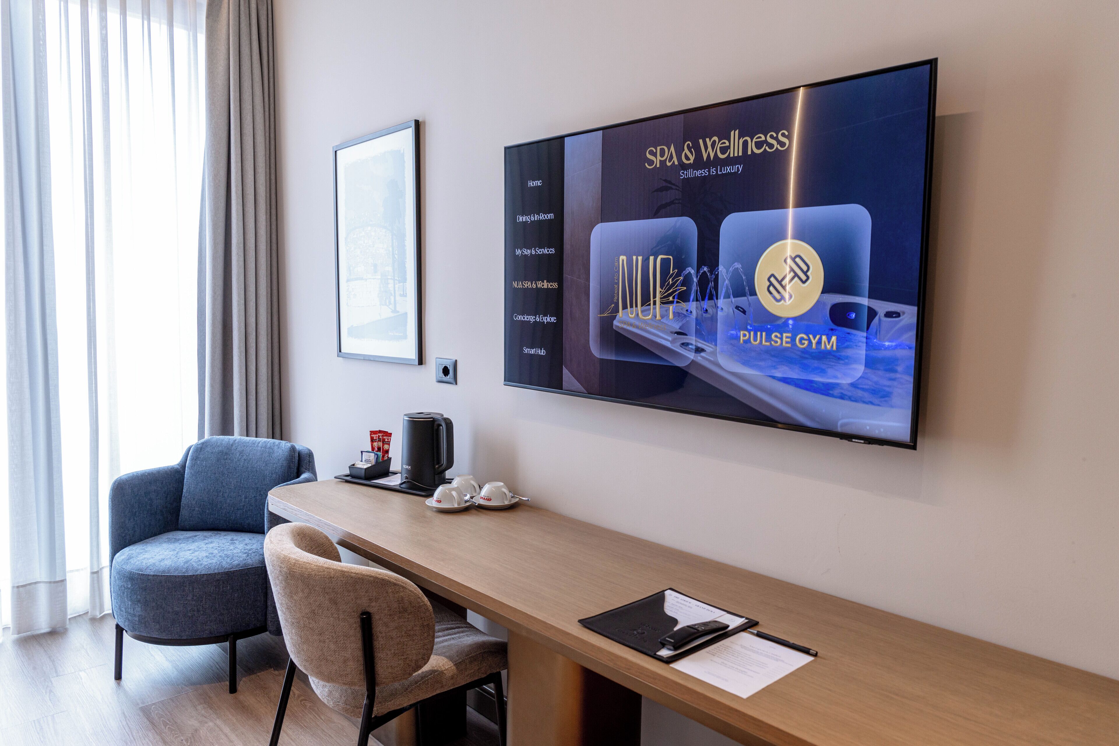 A hotel room with a large TV displaying a spa and wellness advertisement, a wooden desk, a chair, and a coffee maker.