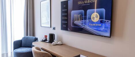 A hotel room with a large TV displaying a spa and wellness advertisement, a wooden desk, a chair, and a coffee maker.