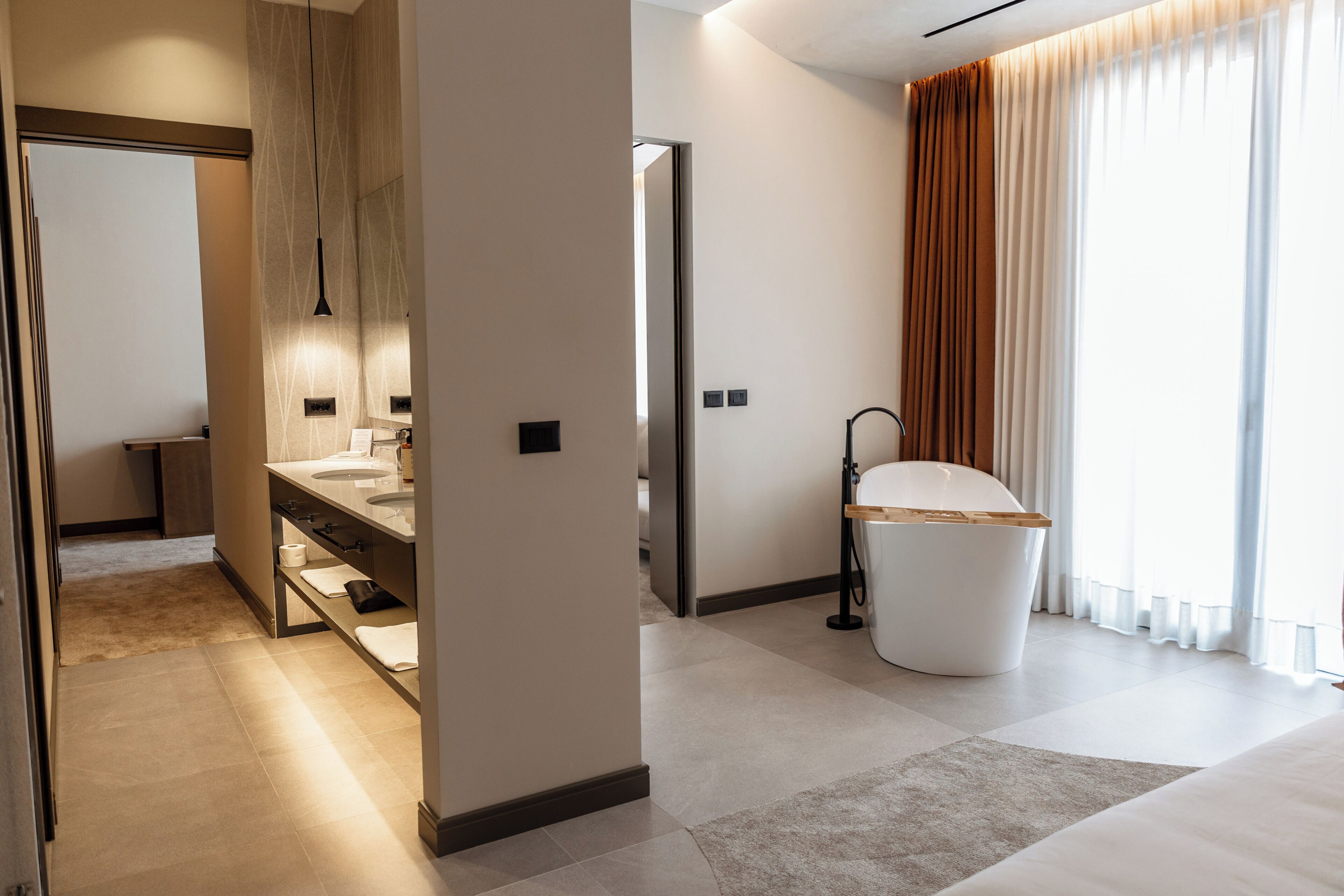 A modern hotel room with a bathroom, a large window with curtains, and a bed.