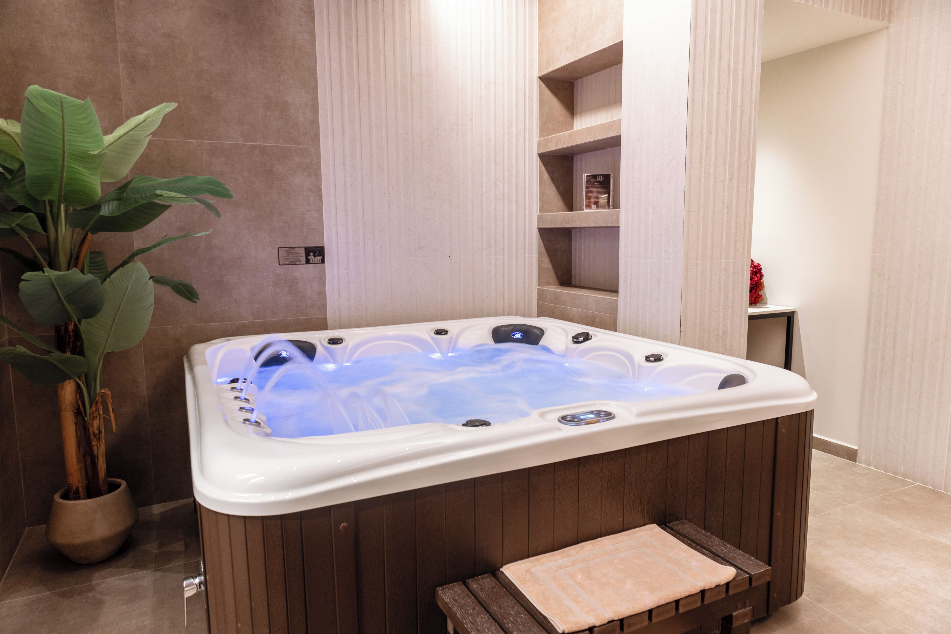 A hot tub in a spa area with a wooden bench, a potted plant, and a tiled wall.