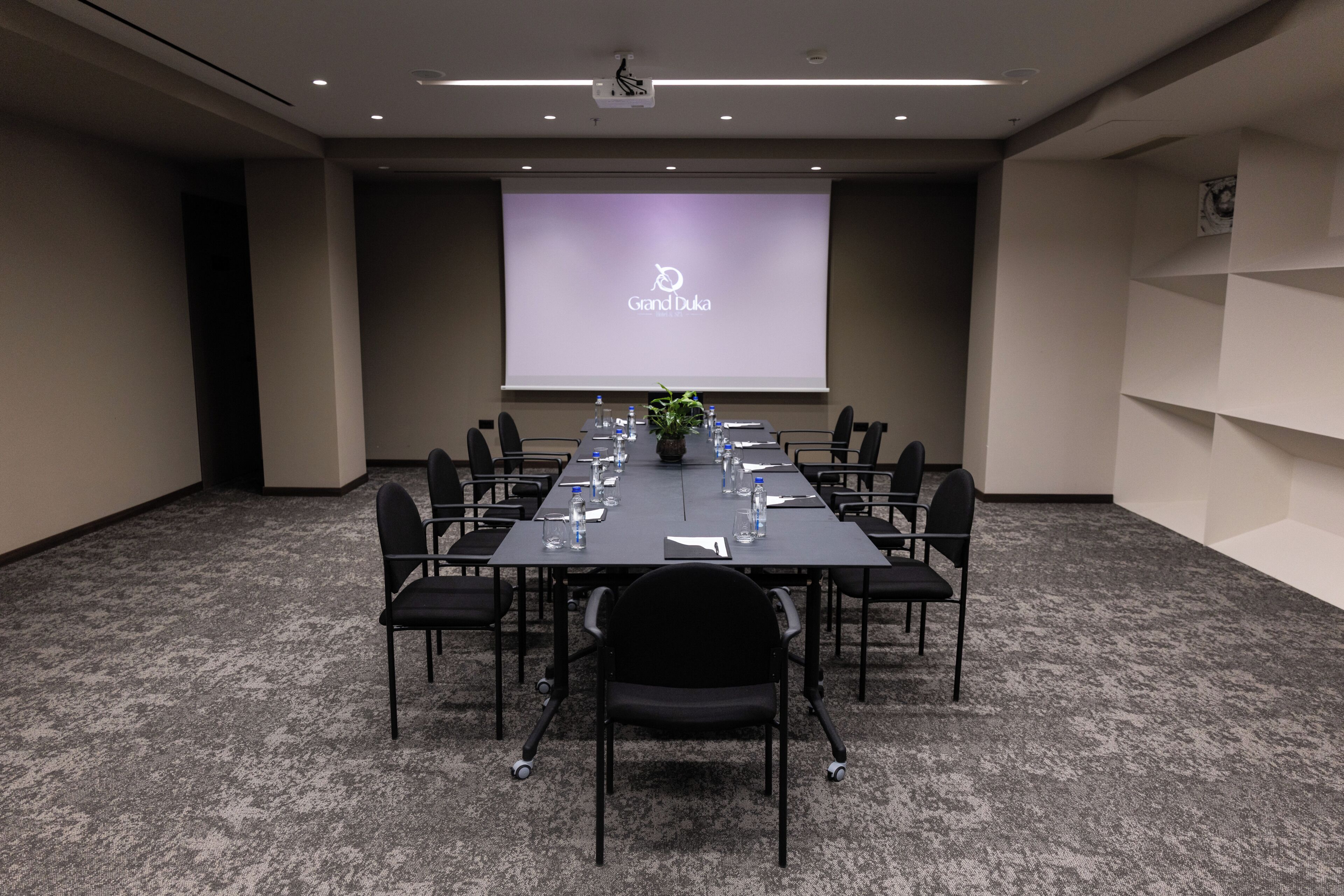 A conference room with a long table, chairs, a projector screen, and a carpeted floor.