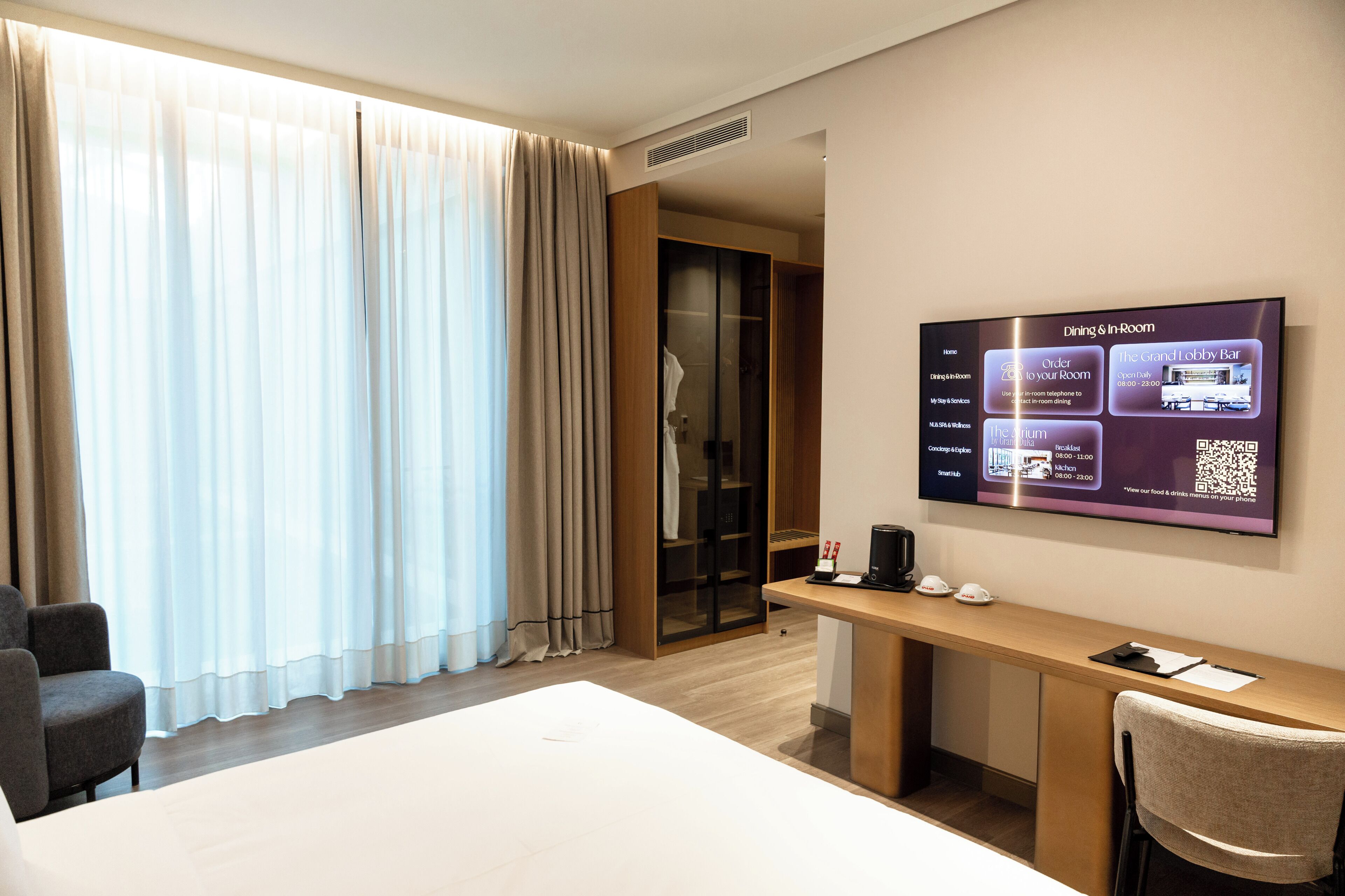 A hotel room with a bed, a desk, a chair, a TV displaying room amenities, and a sliding door closet.