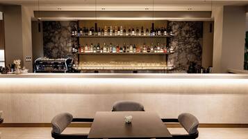 A bar area with a marble wall, multiple shelves of bottles, a coffee machine, and a seating area with two chairs and a table.