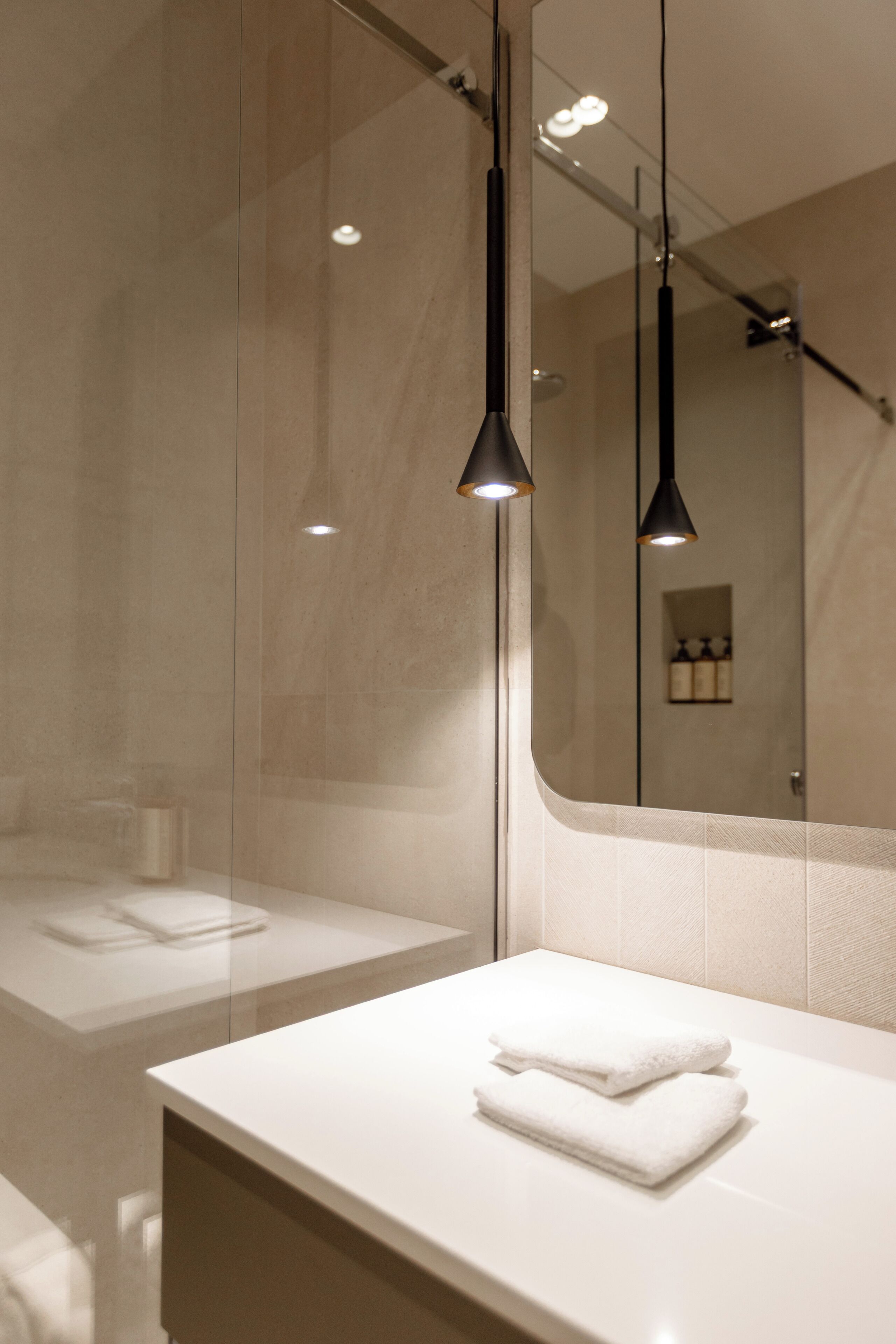 Et moderne badeværelse med glasbrusekabine, et hvidt toiletbord med et håndklæde og hængende lys.