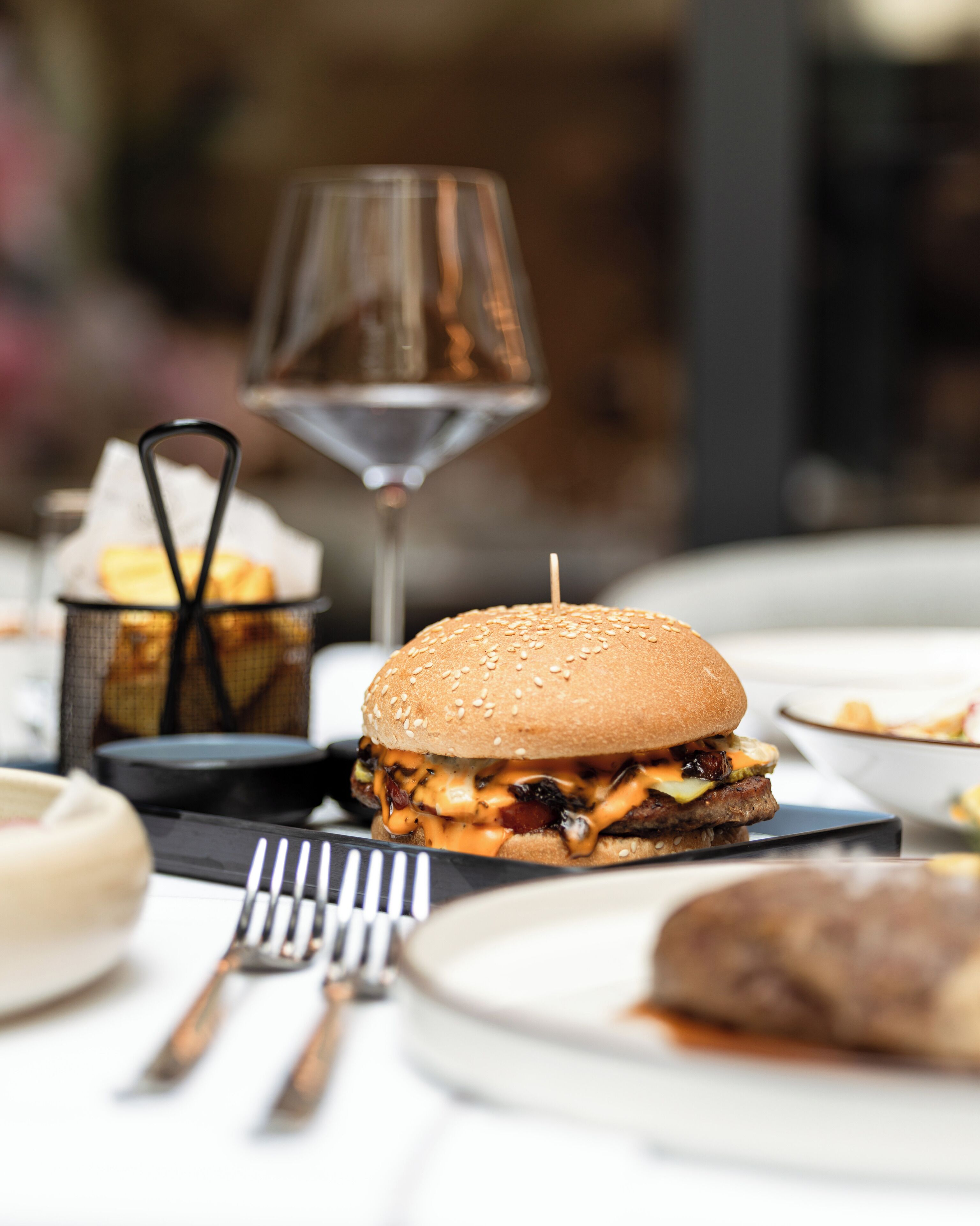 A dining table with a burger, a glass of wine, and utensils.
