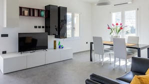 Una sala moderna con un televisor de pantalla plana, muebles blancos, una zona de comedor con mesa y sillas, y ventanas amplias con vistas.