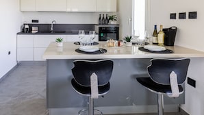 Una cocina moderna con armarios blancos, horno empotrado y una zona de comedor con dos taburetes negros.