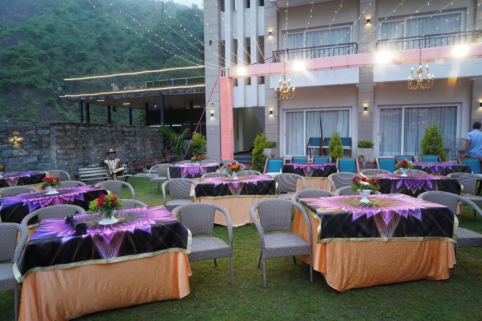 Un área de comedor al aire libre con mesas dispuestas para comensales, rodeada de sillas de mimbre y cubiertas con manteles.