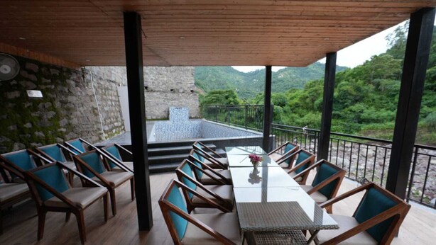 A covered outdoor seating area with a long table and chairs, stone walls, and a view of a mountainous landscape. - Royal suites Chakki Morh (Kasauli)