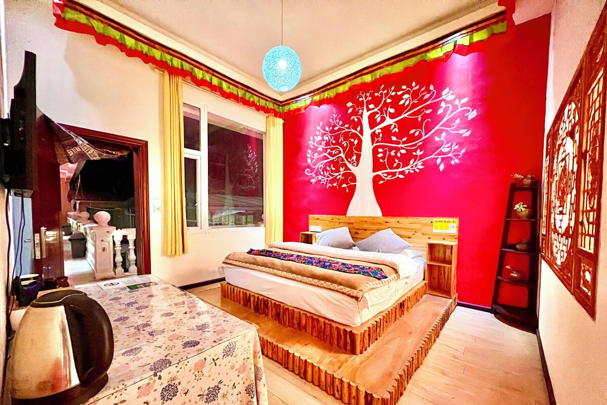 Een slaapkamer met een bed, een houten hoofdbord, een nachtkastje, een lamp, een raam en een rode muur met een muurschildering van een boom.