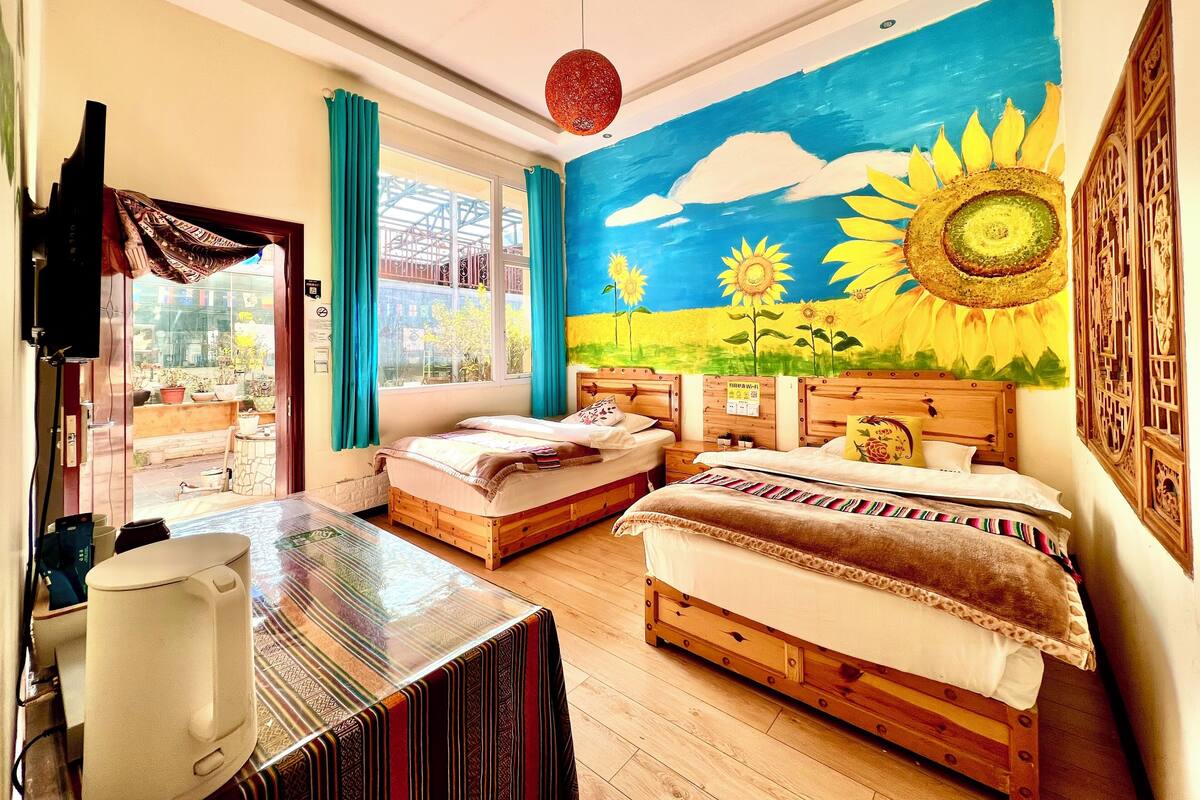 Een kamer met twee bedden, een groot raam met gordijnen, een muurschildering van zonnebloemen en een houten tafel.