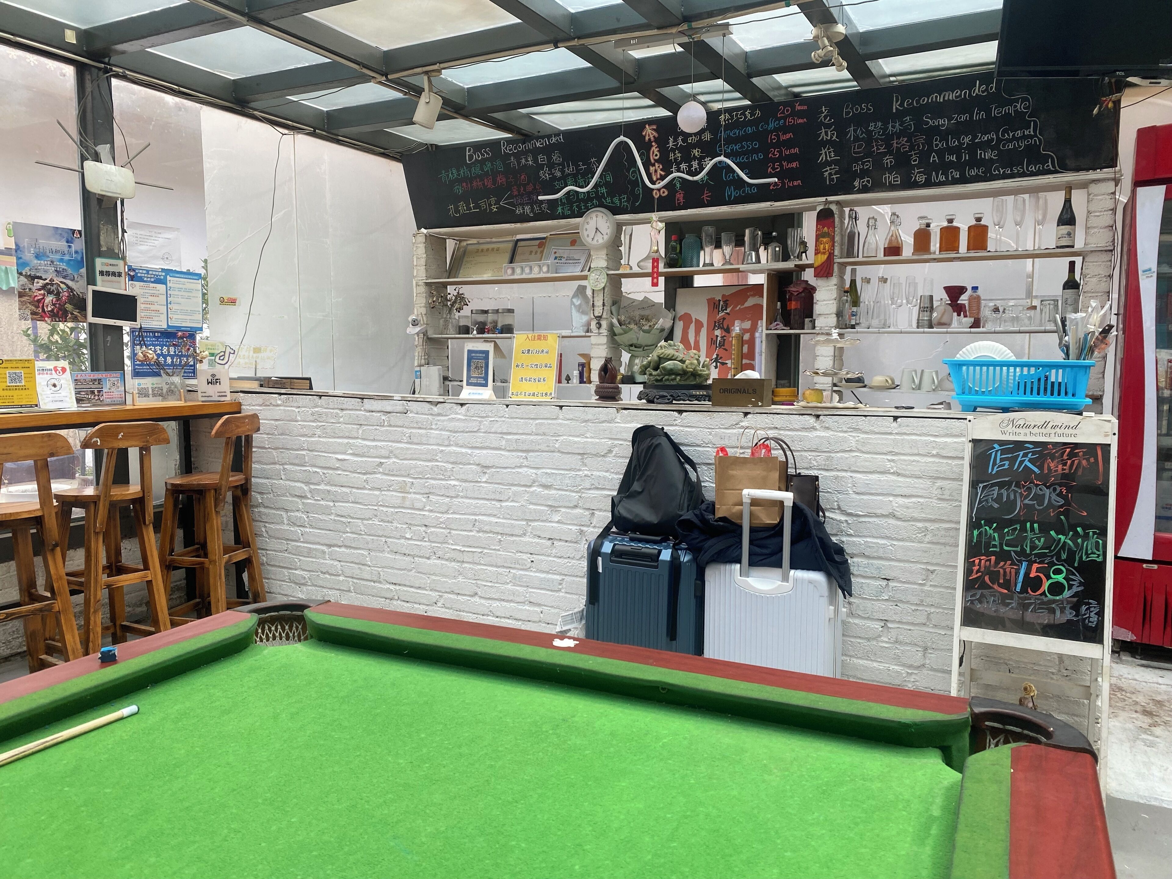 Una mesa de billar con taco, una zona de bar con taburetes, un menú en pizarra y diversas botellas y vasos en estantes.