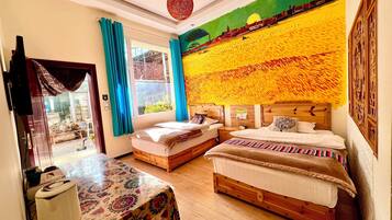 A hotel room with two beds, a table, and a view of a garden through a window.