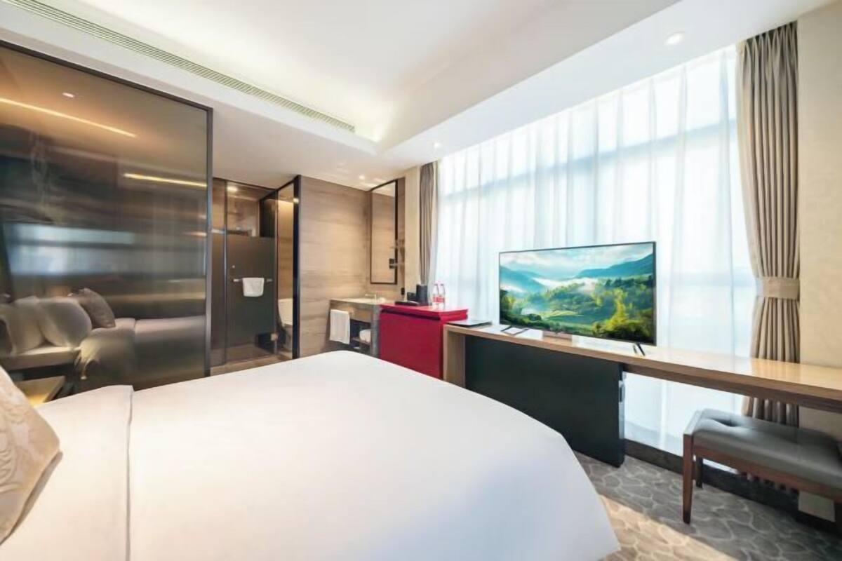 Una habitación de hotel moderna con una cama grande, un escritorio con televisor de pantalla plana, una zona de estar con sofá y un amplio ventanal con cortinas.